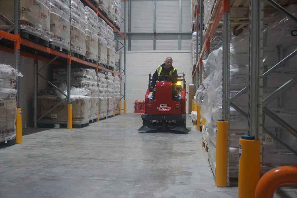 A man sitting on a MAC sweeper cleaning the floor of a warehouse.