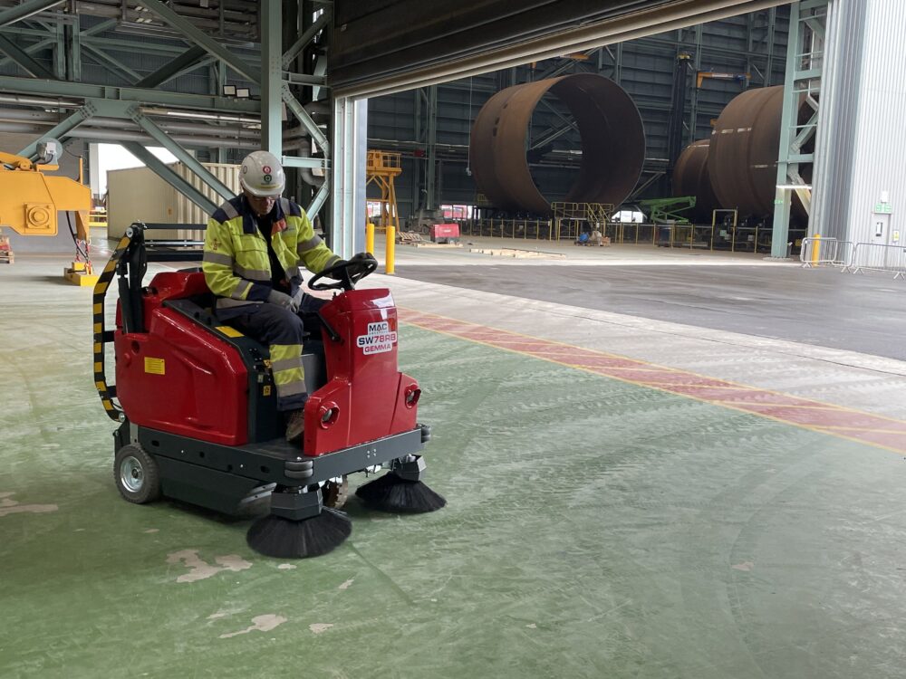 A worker wearing PPC in a warehouse using a MAC ride-on sweeper.